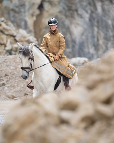 Eine Frau ist mit Helm und Regenreitmantel bekleidet und reitet auf einem weißen Pferd mit grauer Mähne.