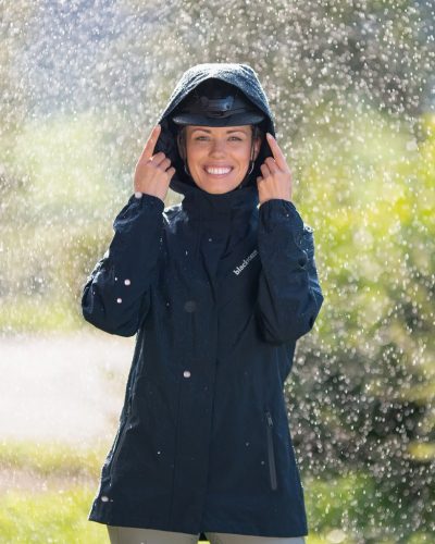 Eine Frau trägt eine Regenjacke zum Reiten und hat die Kapuze über den Helm gezogen. 